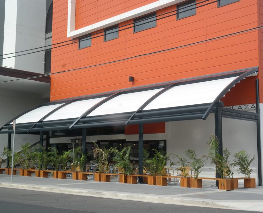 In front of road there are multiple plants on the wooden benches and a Orange Color texture building with the white color roof shade