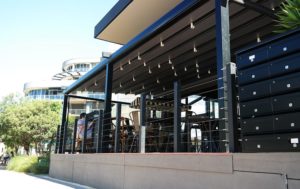 With the buildings and green trees in the background, an outdoor cafeteria with a black metal ceilings, dining tables, and several lockers