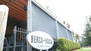 Aluminium sheet interior shutter rolls on the side walls of the café bar with beach house café bar entrance board in front of lovelygarden and bar contains black color chairs.