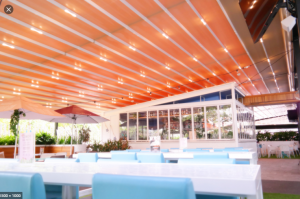 The outdoor dining area of the restaurant and folding roof with lamps and white tables with sky blue color chairs in the restaurant